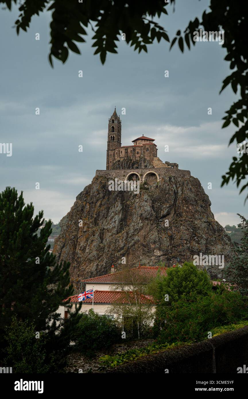 Porträtaufnahme der Kirche Saint Michel auf einem vulkanischen Felsen in Le Puy-en-Velay, eingerahmt von umliegenden Bäumen vor einem bewölkten Himmel. Stockfoto