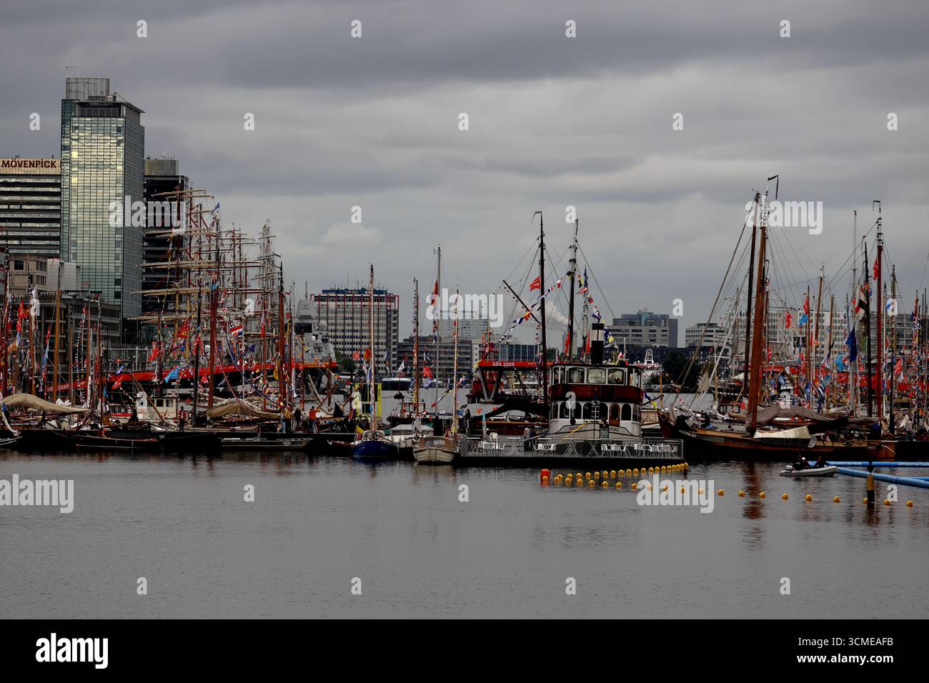 Eine gemischte Flotte von Segelschiffen und klassischen Booten säumt die IJ für Sail 2025 in Amsterdam, Niederlande, Takelage und Flaggen unter einem bewölkten Himmel. Stockfoto