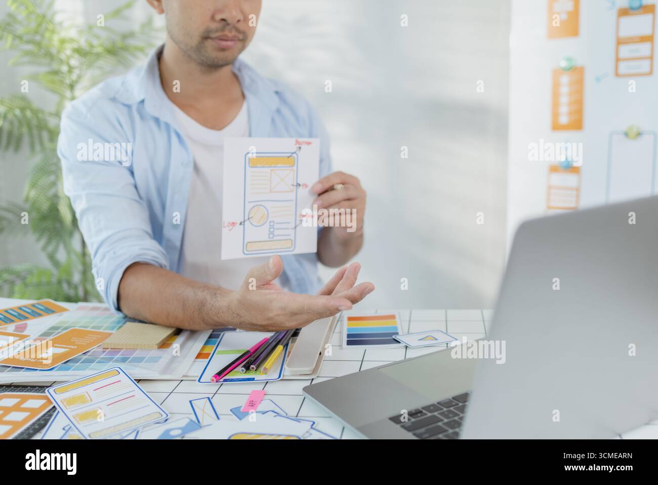 Ein fokussierter Designer arbeitet an UX/UI-Designideen und analysiert Farbpaletten und Schnittstellenstrukturen, die auf einer Platine fixiert sind. Die Szene spiegelt Kreativität wider Stockfoto