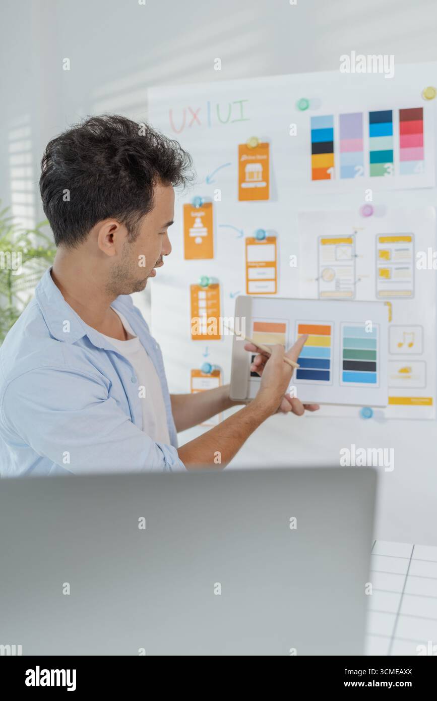 Ein fokussierter Designer arbeitet an UX/UI-Designideen und analysiert Farbpaletten und Schnittstellenstrukturen, die auf einer Platine fixiert sind. Die Szene spiegelt Kreativität wider Stockfoto