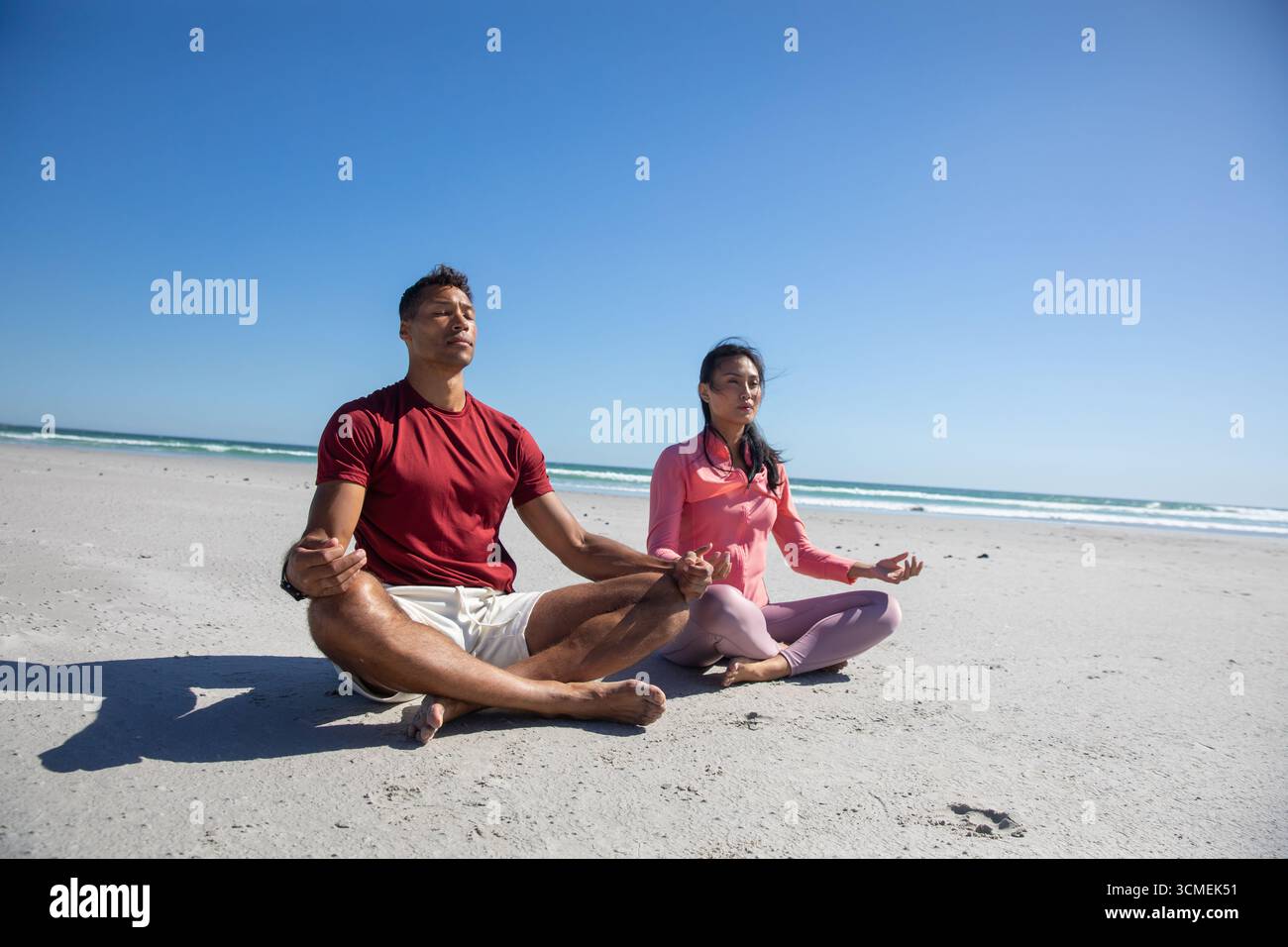 Diverse Paare üben Meditation im Kreuz am Strand am Meer unter klarem Himmel Stockfoto