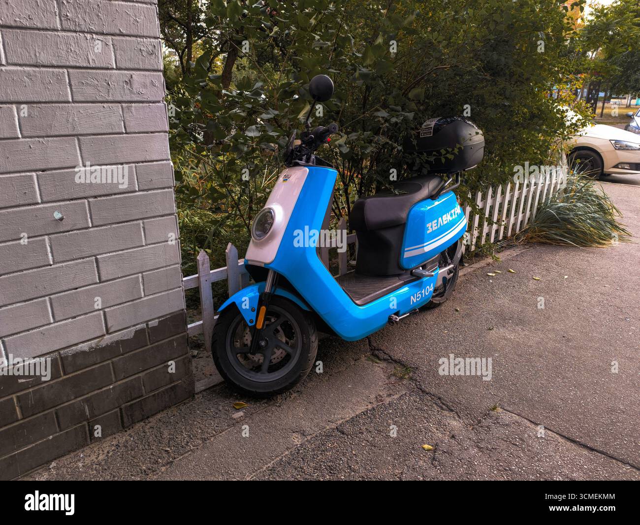 Geparkter blauer Elektroroller mit Helm in der Nähe von Ziegelmauern und Büschen. Stockfoto