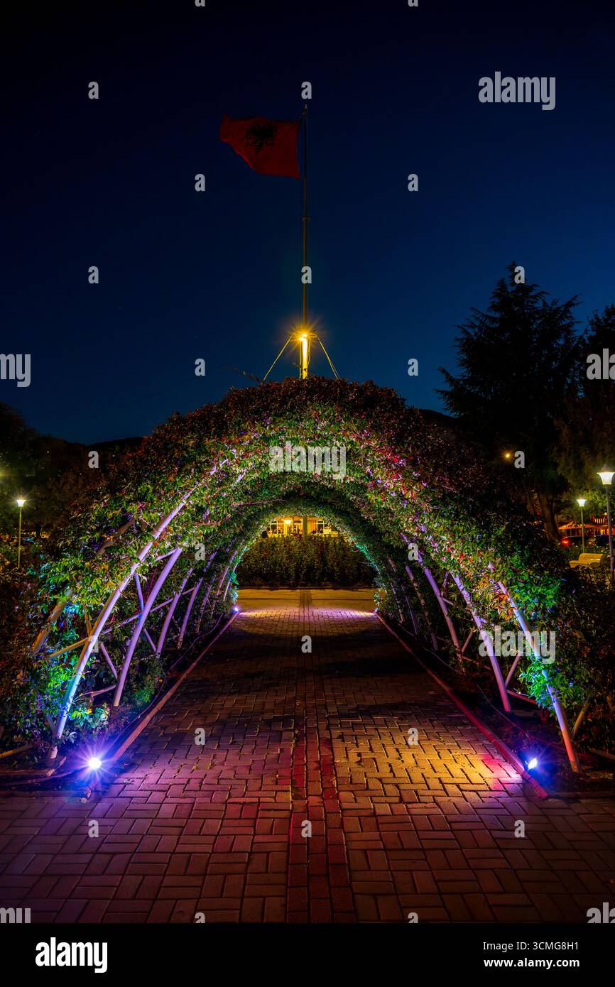 Beleuchteter Floral Bogenway und albanische Nationalflagge bei Nacht – lebendiger Weg in einem Stadtpark Stockfoto