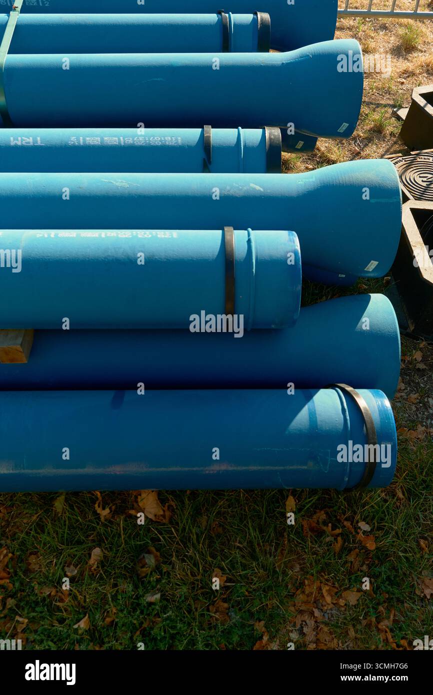 Vertikales Foto von ordentlich gestapelten blauen Industrierohren und -Röhren auf grünem Gras, verstreut mit gelben Herbstblättern. Helles Tageslicht hebt die Textur hervor Stockfoto