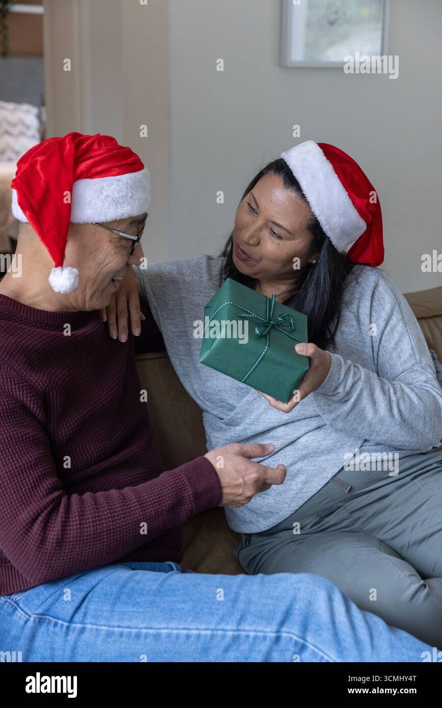 Asiatische Familie tauscht grün verpacktes Geschenk auf Wohnzimmer Sofa mit weihnachtsmützen aus Stockfoto