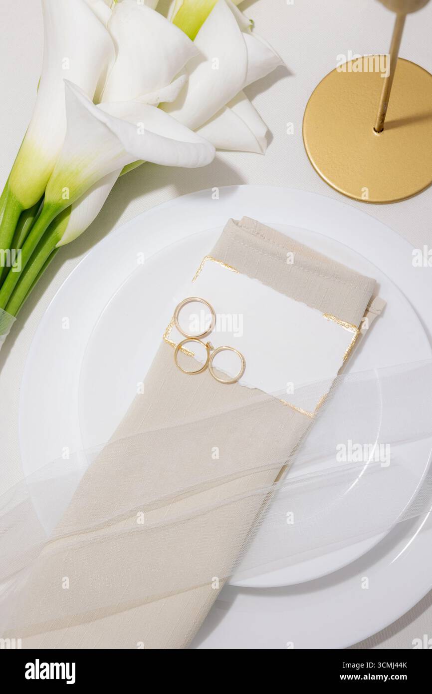 Setzen Sie eine romantische Atmosphäre mit wunderschönen weißen Blumen, zartem Serviettendekor und goldenen Ringen auf einem elegant gestalteten Tisch. Stockfoto