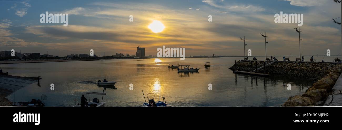 Kleine Fischerboote werden auf ruhigen Gewässern bei Sonnenuntergang in der Nähe des Hafens in Kuwait City, Kuwait, gesehen, mit modernen Gebäuden in der Ferne unter einem cl Stockfoto