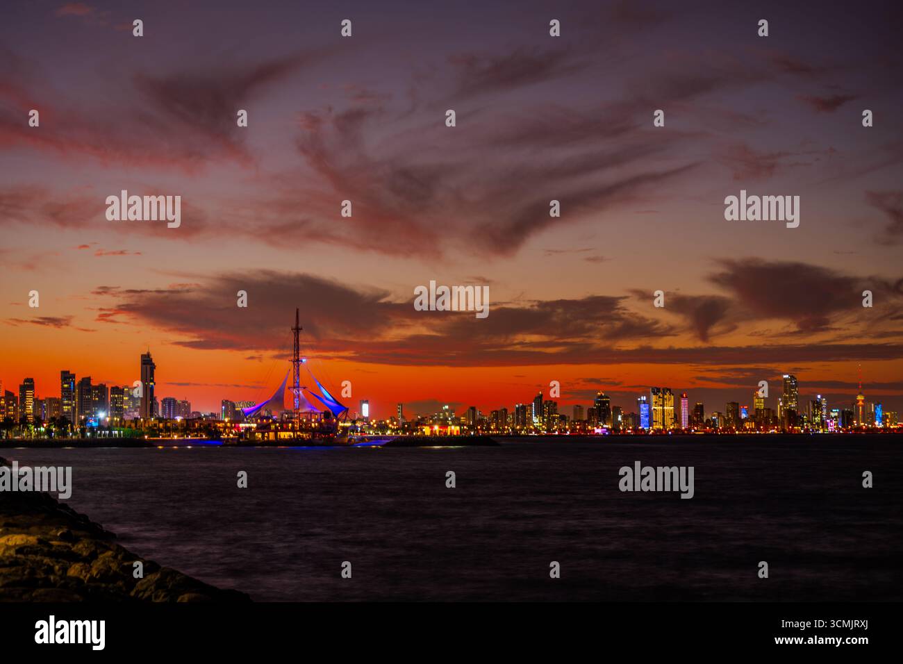 Atemberaubender Blick bei Dämmerung auf die beleuchtete Skyline von Kuwait City, mit dem Marina Crescent Sailing Tower, der vor einem pulsierenden Sonnenuntergang über der Ara leuchtet Stockfoto