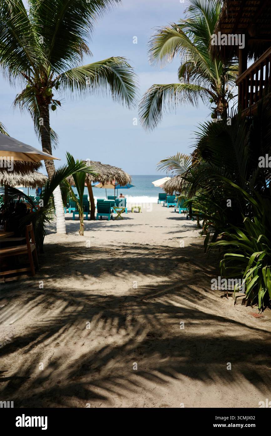 Ein sandiger Pfad durch Palmen führt zum Strand La Punta in Puerto Escondido mit Palapas und türkisfarbenen Stühlen, die gegen die pazifische Brandung stehen. Stockfoto