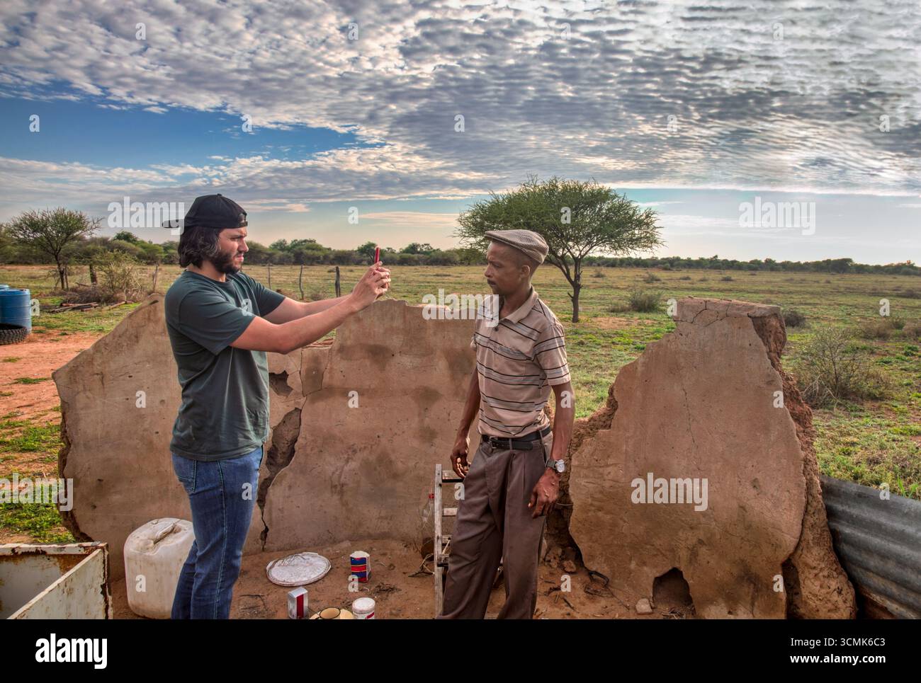 Freiwilliger Sozialarbeiter bei einer gemeinnützigen Organisation, der Fotos von einem afrikanischen Dorfmenschen vor der zerstörten Mauer macht, im Hof bei Sonnenuntergang. Stockfoto