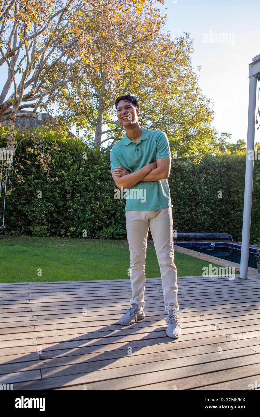 Mann Anfang 20, der mit gefalteten Armen auf Holzdeck in der Nähe von Hecke und Poolabdeckung stand Stockfoto