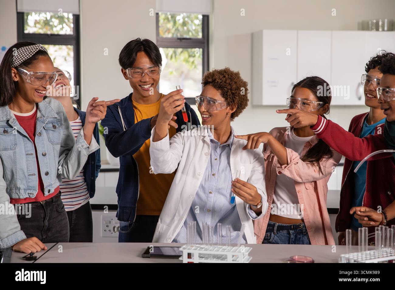 Afroamerikanische Frau im Labormantel, die ein rotes Teströhrchen hält, das Jugendliche Schüler auf dem Labortisch führt Stockfoto