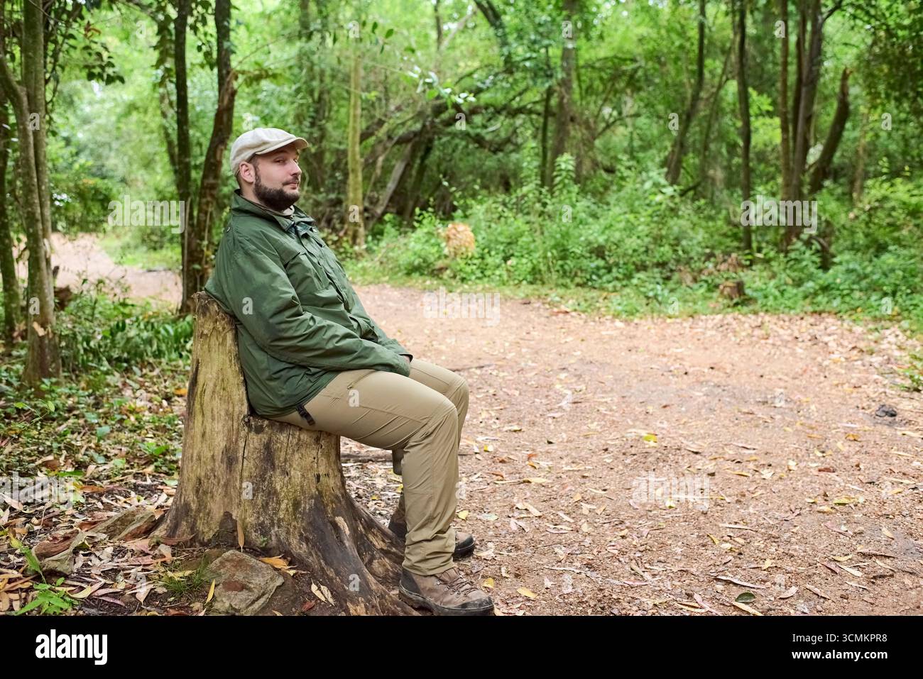 Junger hispanischer Mann, der auf einem Baumstumpf sitzt, als Sitz neben einem Wanderweg in einer natürlichen Umgebung, El Palmar Nationalpark, Entre Rios, Argentinien. Stockfoto