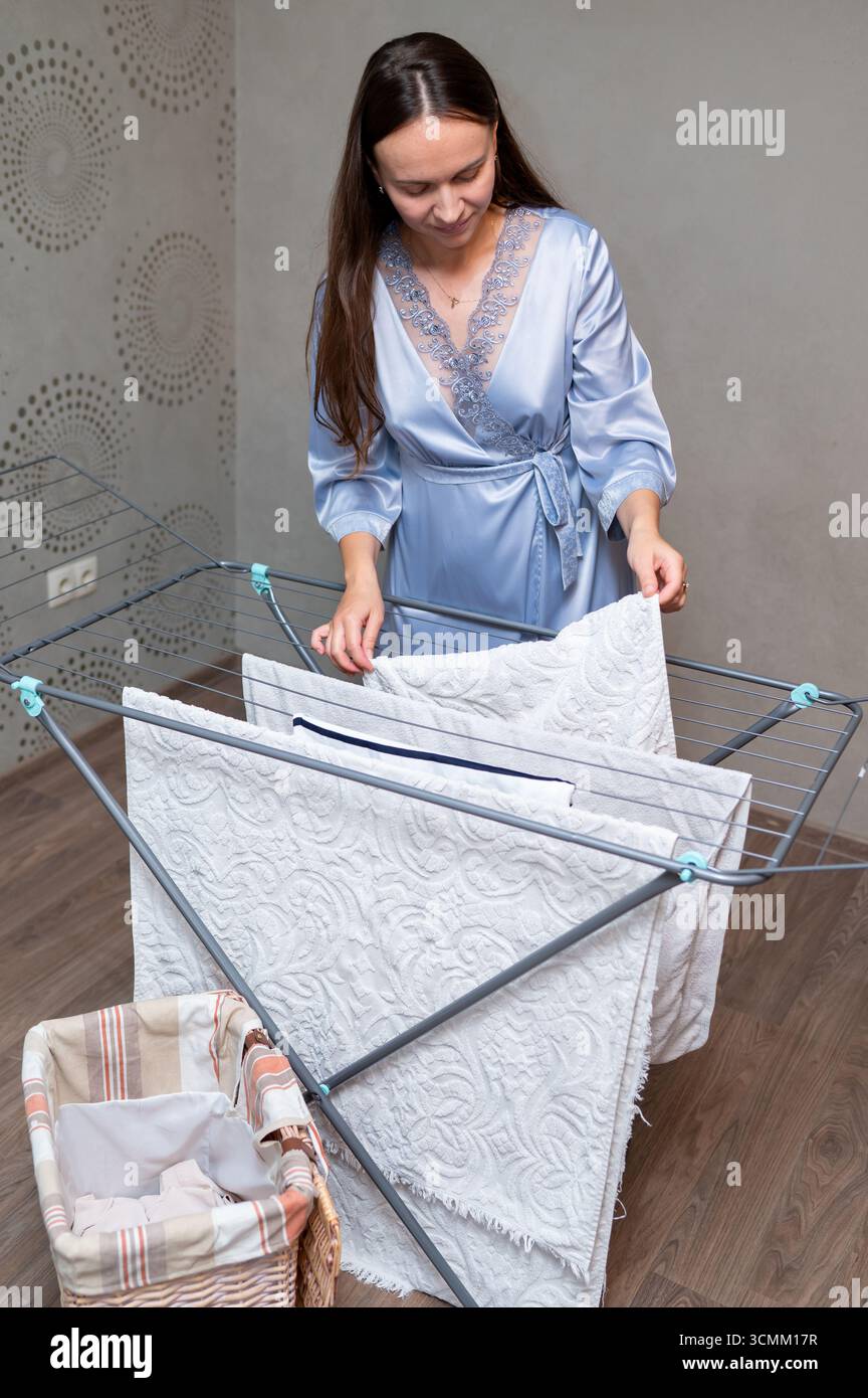 Elegante Hausfrau in Seidenmantel mit lockeren Haaren, die saubere Kleidung auf einem Wäscheständer hängen, Konzept der Hausarbeit und des häuslichen Lebensstils Stockfoto