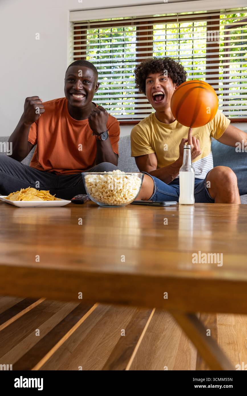 Verschiedene männliche Freunde jubeln und drehen Basketball auf der Couch zu Hause mit Popcorn, Chips, Drink Stockfoto