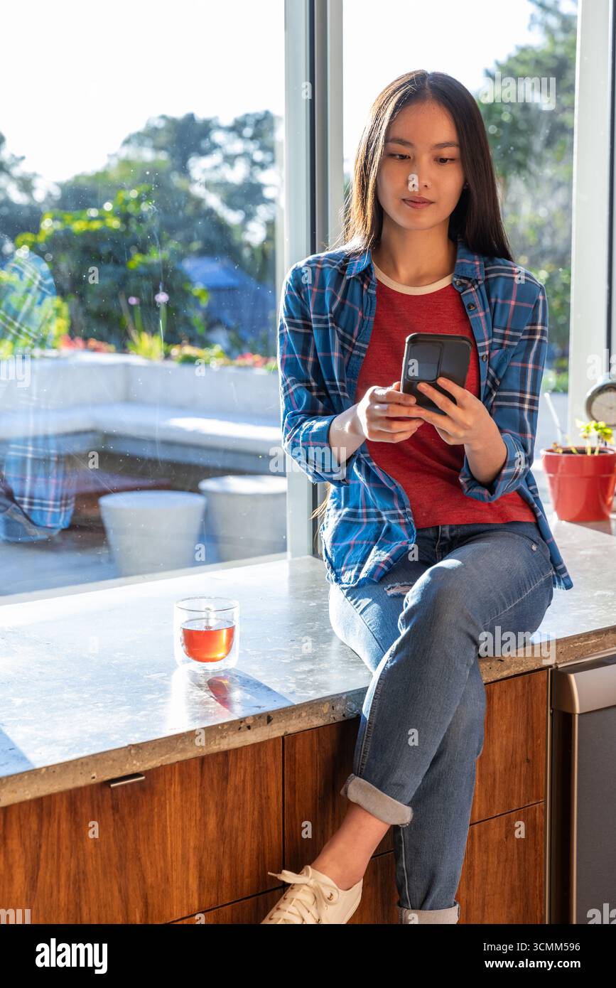 Asiatische Frau, die auf Marmortheke am Fenster sitzt und Smartphone mit Glasbecher hält Stockfoto