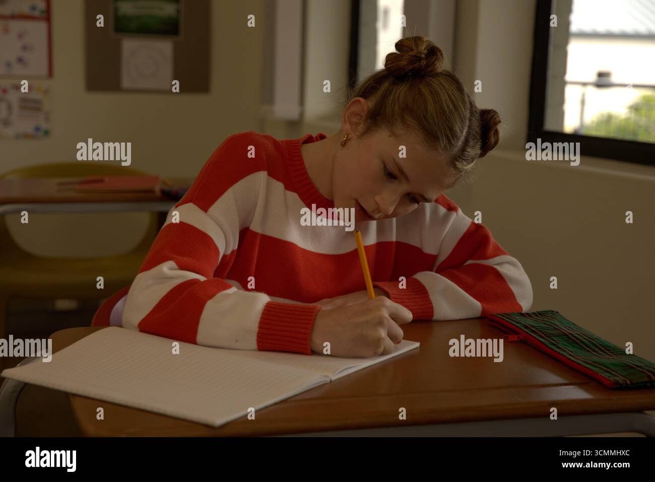 Mädchen in gestreiftem Pullover, das am Schultisch sitzt und in Notizbuch mit gelbem Bleistift schreibt Stockfoto