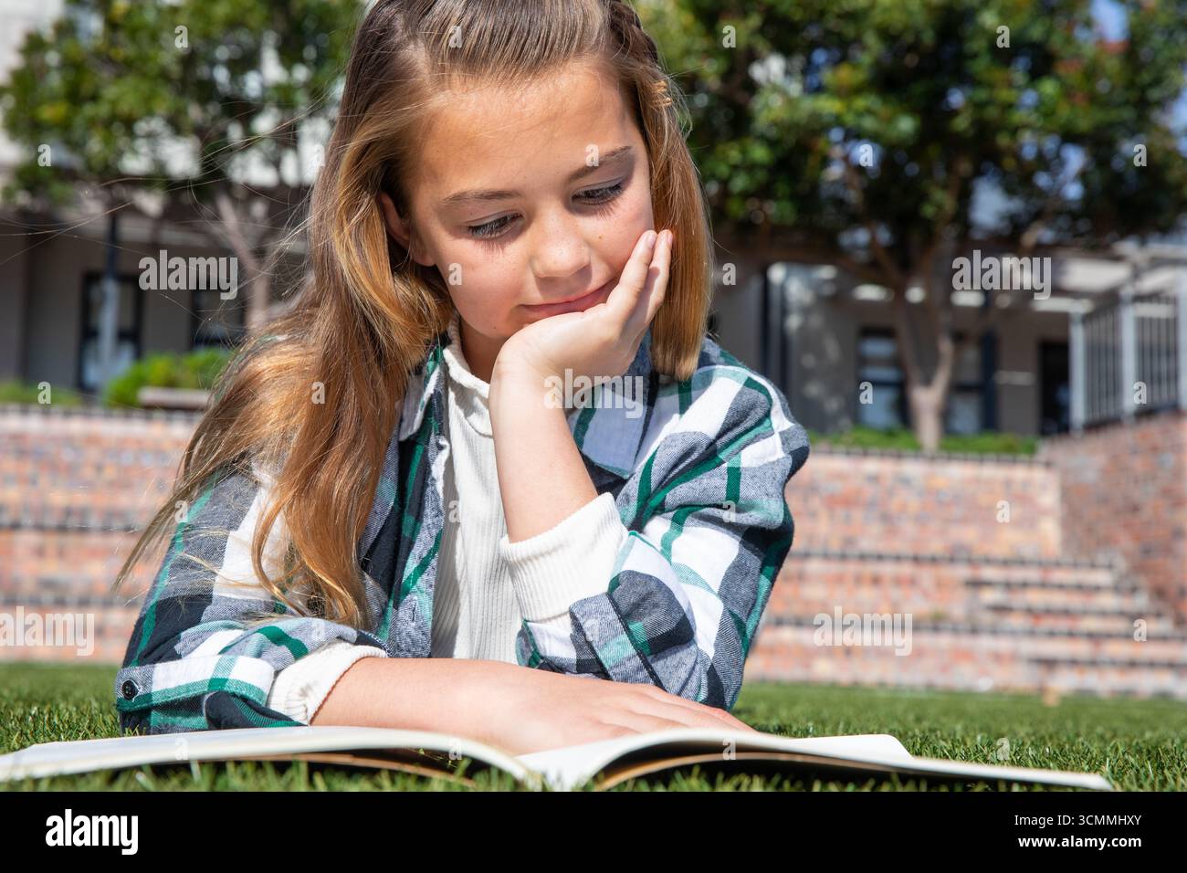 Mädchen, das auf Gras liegt und Buch liest, im Schulhof neben gemauerten Stufen unter sonnigem Himmel Stockfoto