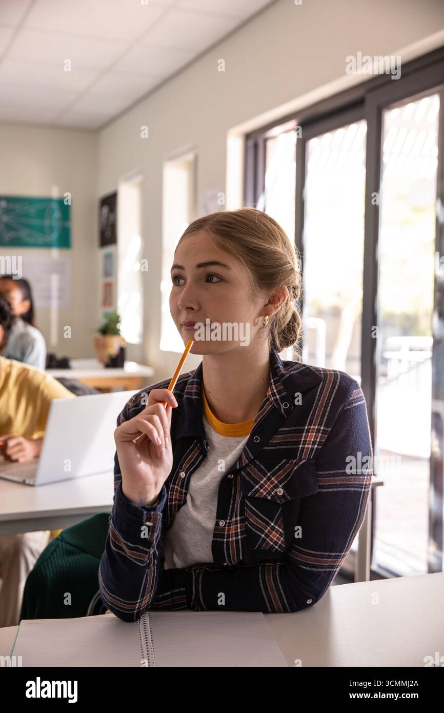 Verschiedene Klassenkameraden im Klassenzimmer halten Bleistift bis Kinn über Notizbuch, die am Laptop tippen Stockfoto