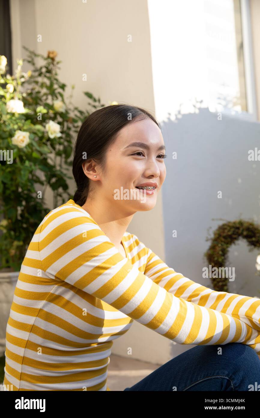 Asiatische Frau in ihren Zwanzigern, die auf dem Balkon sitzt, mit einer Rose, einem Kranz, Fensterläden, lächelnd Stockfoto