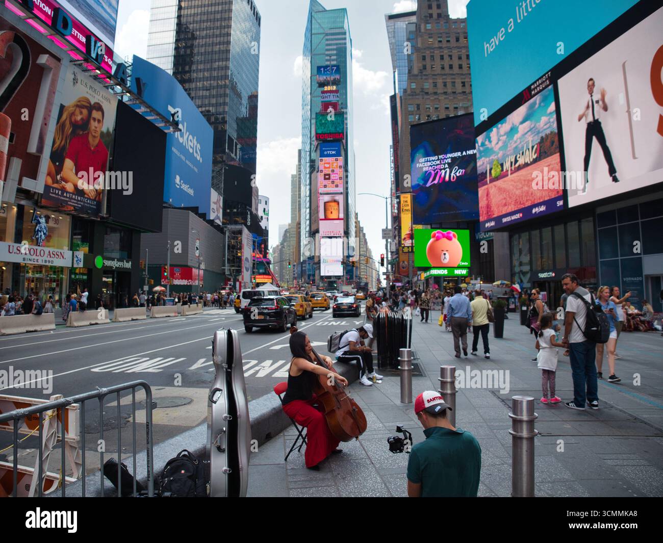 Times Square Street Performer – Eine Frau spielt ein Cello auf dem Bürgersteig am Times Square, New York City, während ein Mann sie filmt, umgeben von riesigen Billboarden Stockfoto