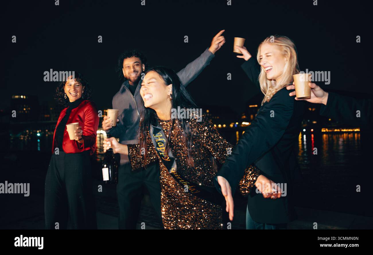 Eine Gruppe fröhlicher Freunde feiert gemeinsam im Freien am Wasser, genießt Drinks und lacht unter den Lichtern der Stadt. Stockfoto