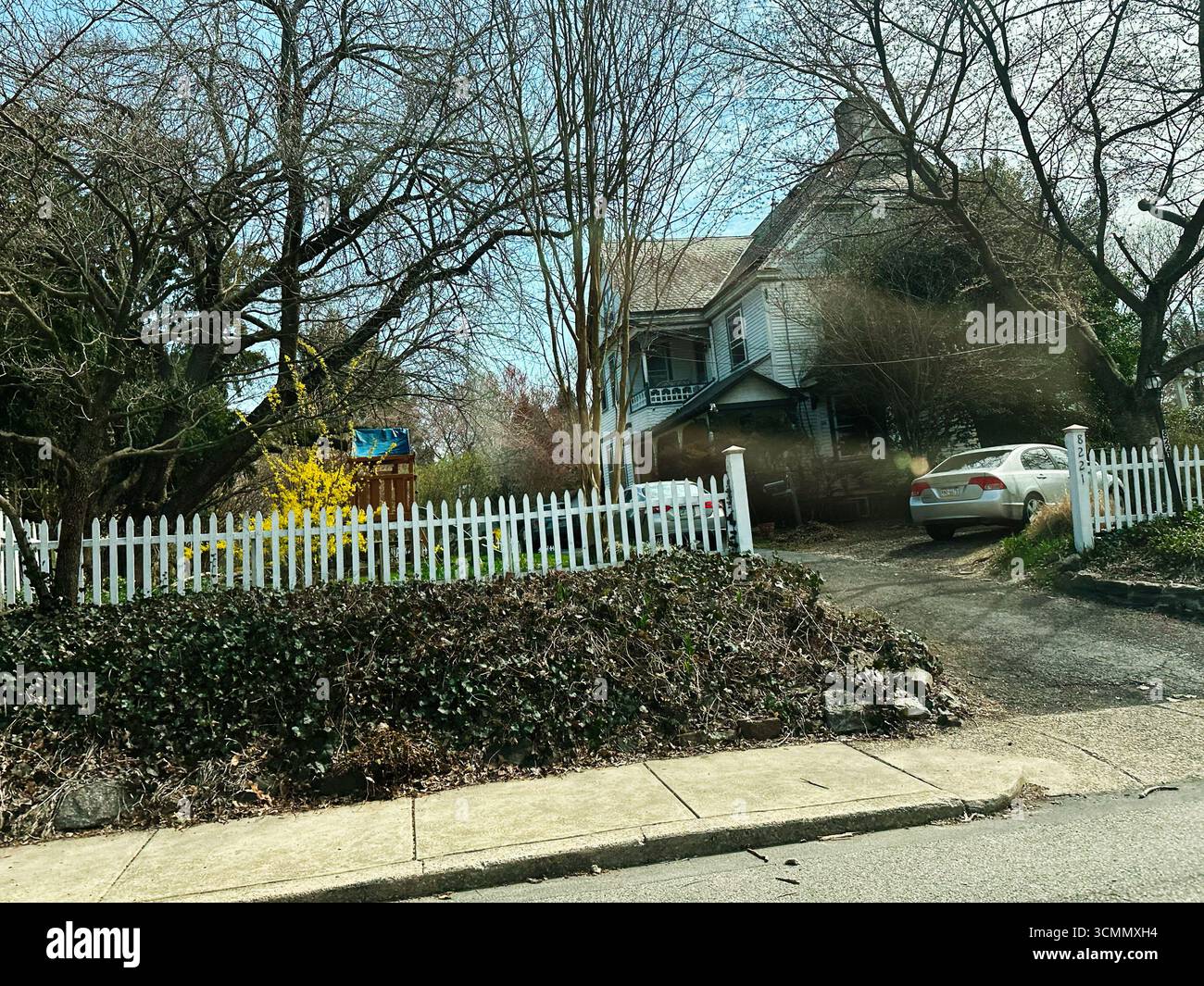 Jenkintown, Pennsylvania, USA - 01. April 2025: Rustikales Haus, umgeben von blühenden Bäumen, einem weißen Streifenzaun und einem Auto. Stockfoto