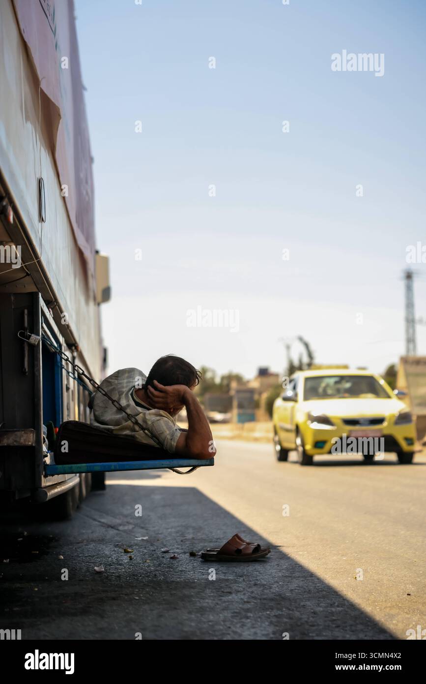 Vorübergehende Pause für einen LKW-Fahrer Ein Mann liegt auf einem provisorischen Bett unter einem geparkten LKW, um der Sommerhitze zu entkommen, an einem Straßenrand in Damaskus. Angesichts schwieriger wirtschaftlicher Bedingungen nach dem Krieg passen sich viele Syrer täglich den rauen Lebensumständen an. Damaskus Syrien Copyright: XMohammadxBashirxAldaherx Stockfoto