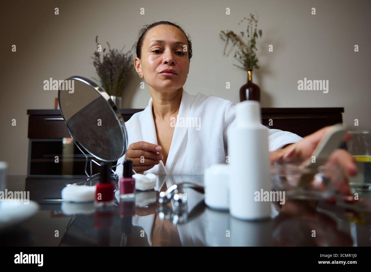 Eine Frau in einem weißen Gewand konzentriert sich auf ihre Selbstpflegeroutine mit verschiedenen Hautpflegeprodukten. Sie steht an einem Schminktisch und betont Wellness und Stockfoto