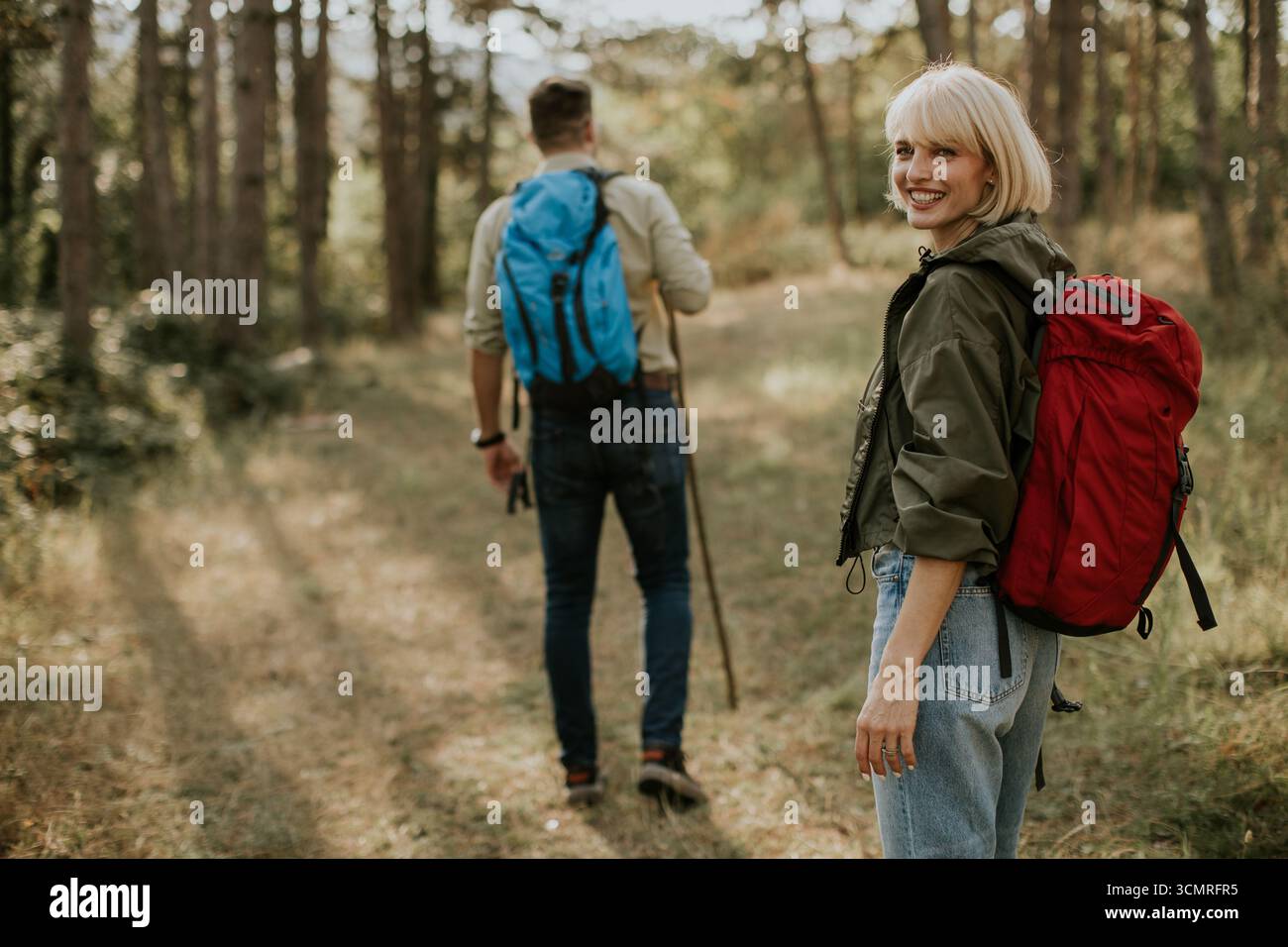 Zwei Wanderer erkunden an einem sonnigen Tag ein Waldgebiet, lächeln und genießen die Natur. Eine Person trägt eine grüne Jacke mit einem roten Rucksack, die andere Person Stockfoto