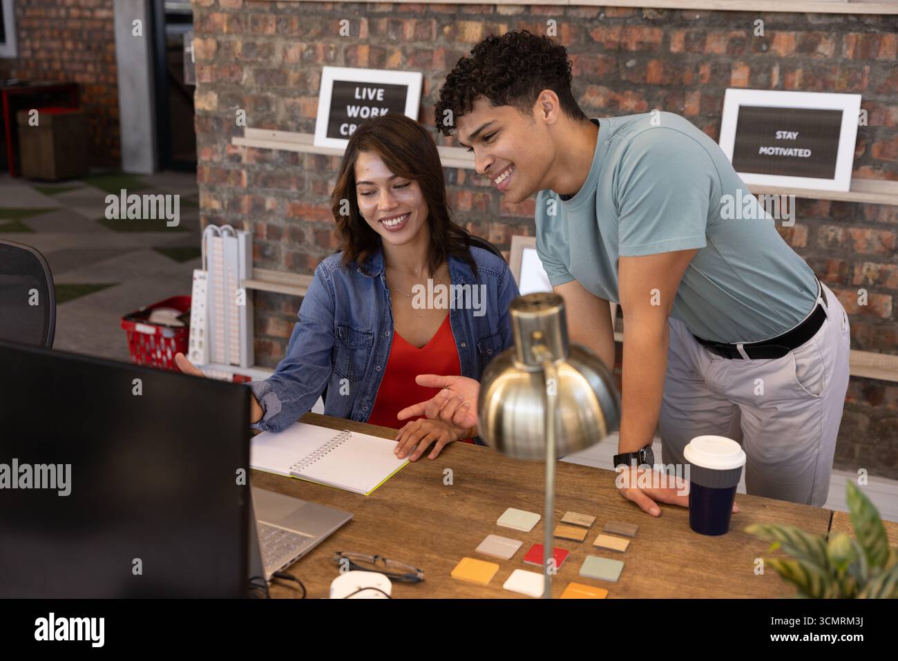 Verschiedene Kollegen arbeiten am Schreibtisch mit Laptops und Notebook-Farbmustern gegen die freiliegende Ziegelwand zusammen Stockfoto