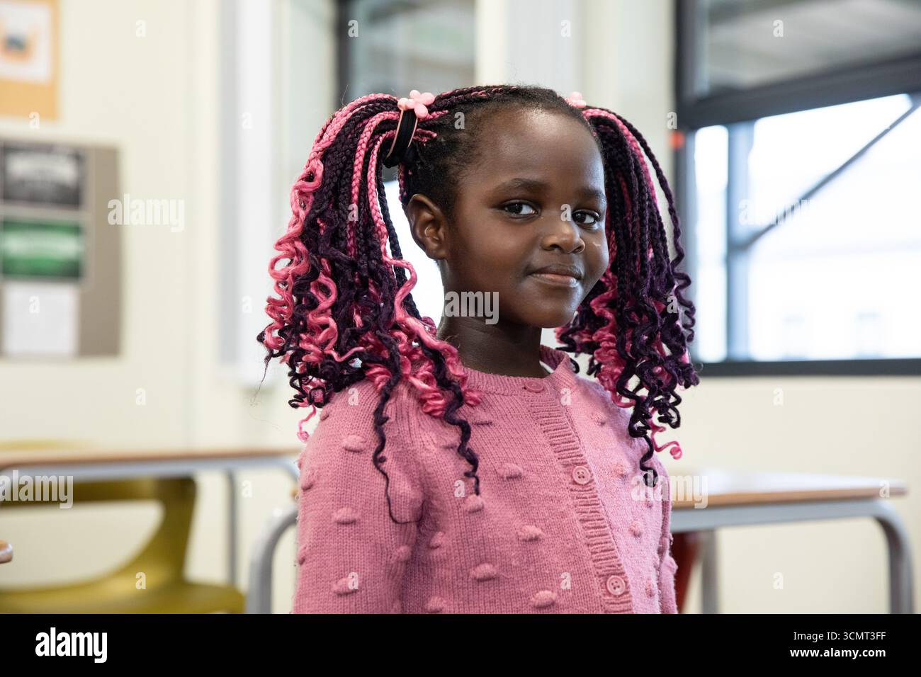 afroamerikanisches Mädchen, das im Klassenzimmer neben Schreibtischen und Stühlen in rosa Pullover steht Stockfoto