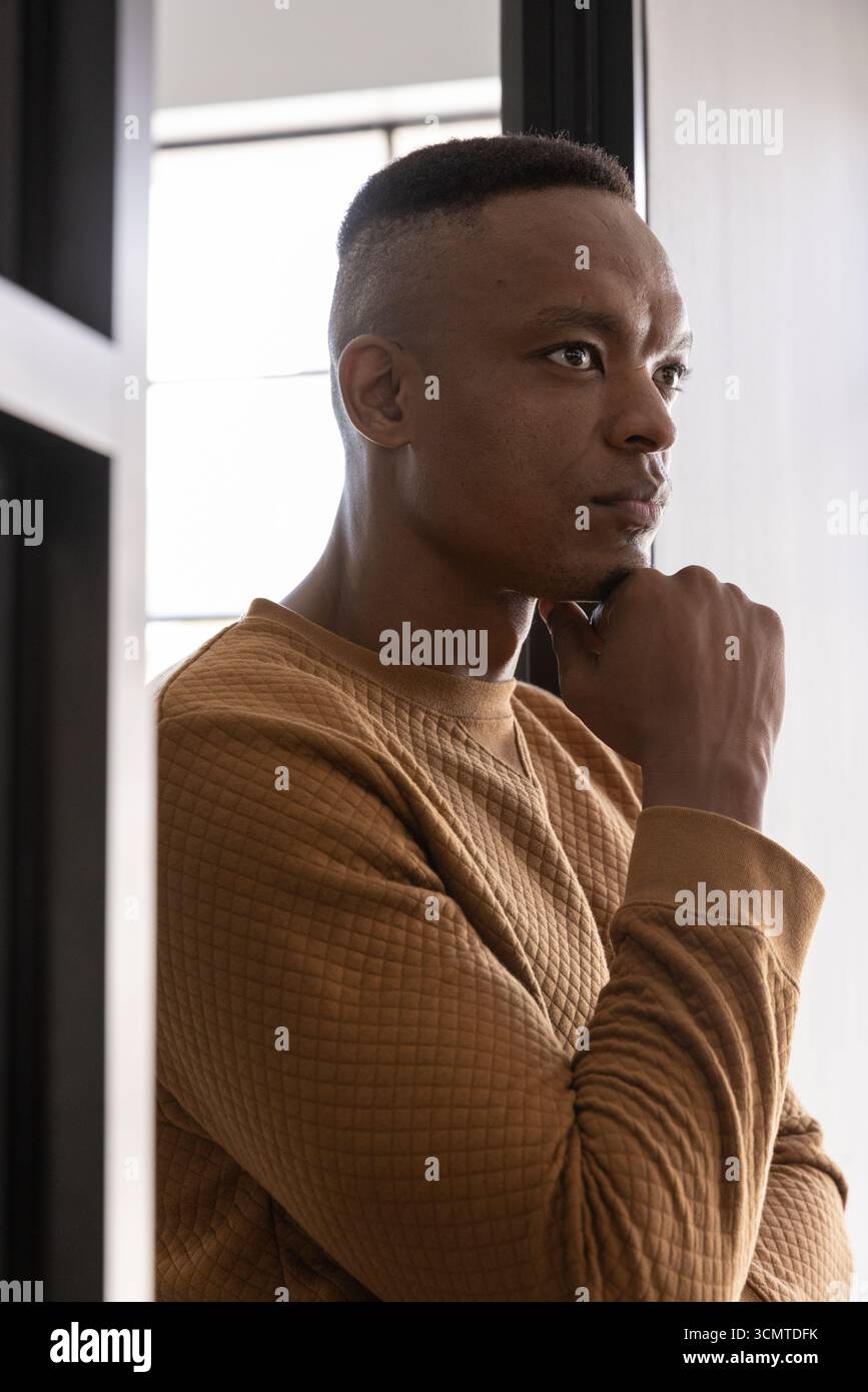 afroamerikaner ruht das Kinn und schaut durch das Fenster im Zimmer mit braunem Sweatshirt Stockfoto