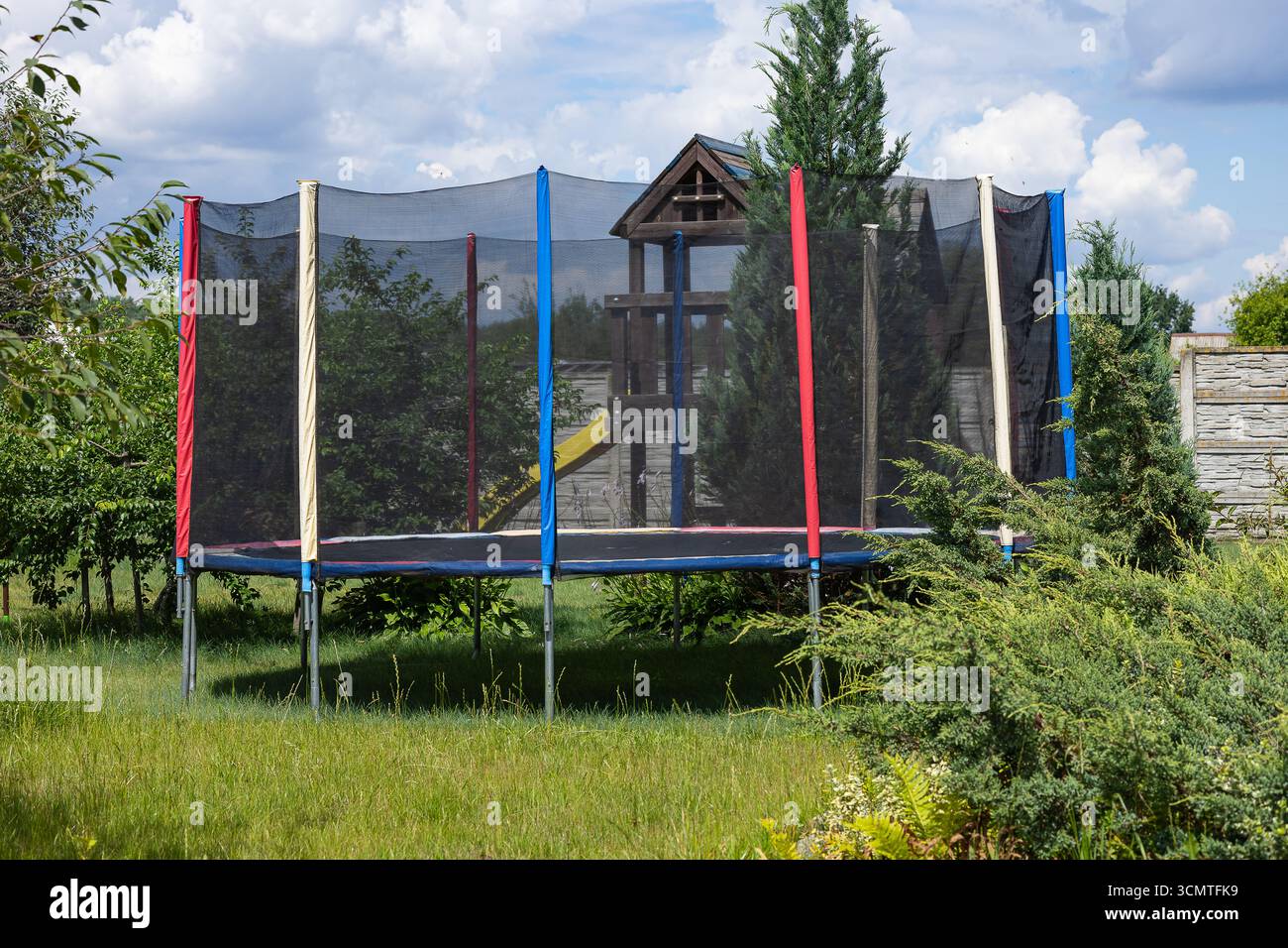 Farbenfrohes Trampolin mit Sicherheitsnetz auf grünem Gras im Garten im Hinterhof, umgeben von Bäumen und Büschen. Outdoor-Aktivitäten und Sportausrüstung Stockfoto