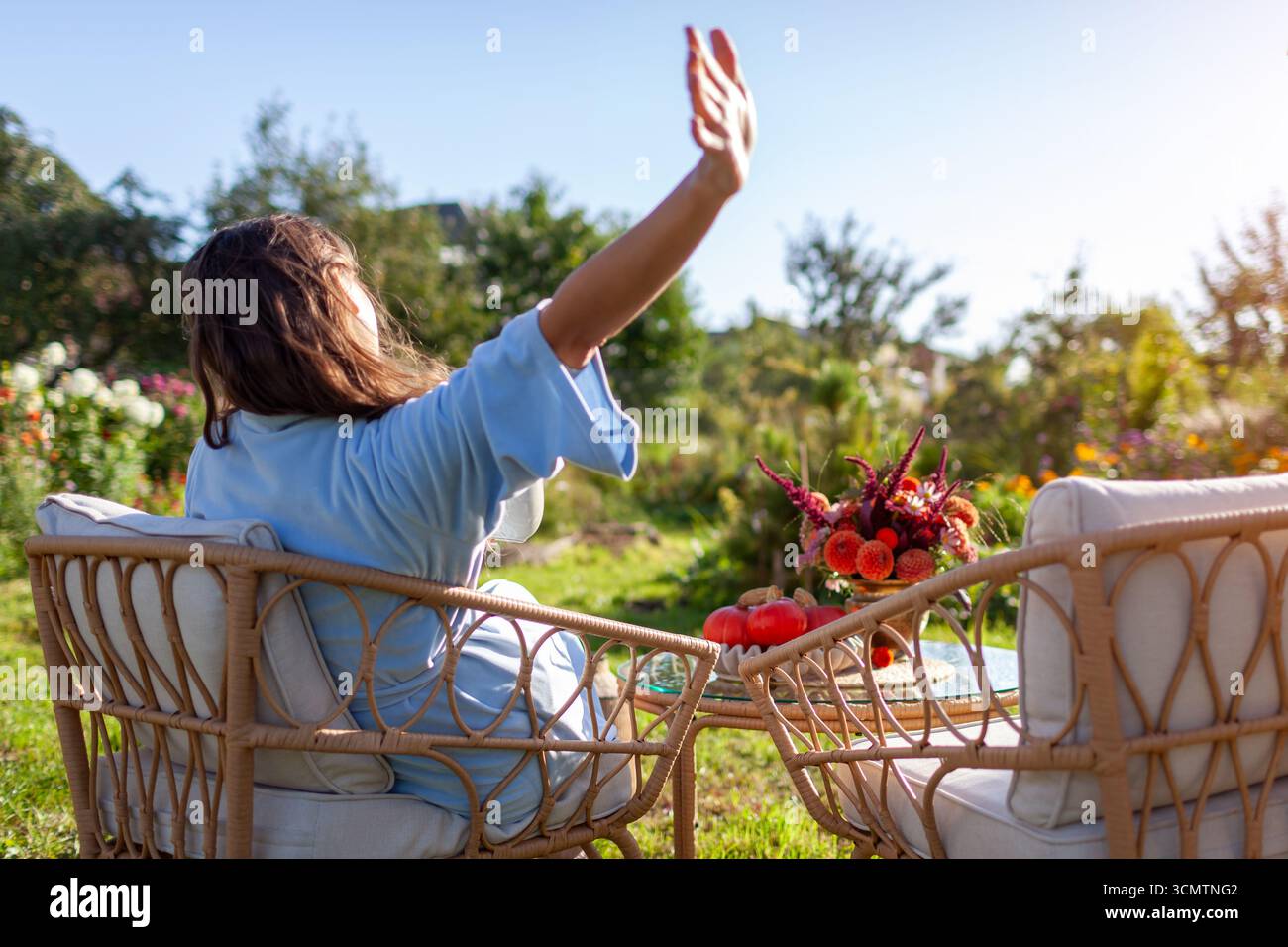 Rückansicht einer Frau, die sich morgens im Pyjama ausdehnt und sich auf Terrassenmöbeln im Herbstgarten entspannt. Kaffee zum Frühstück Stockfoto