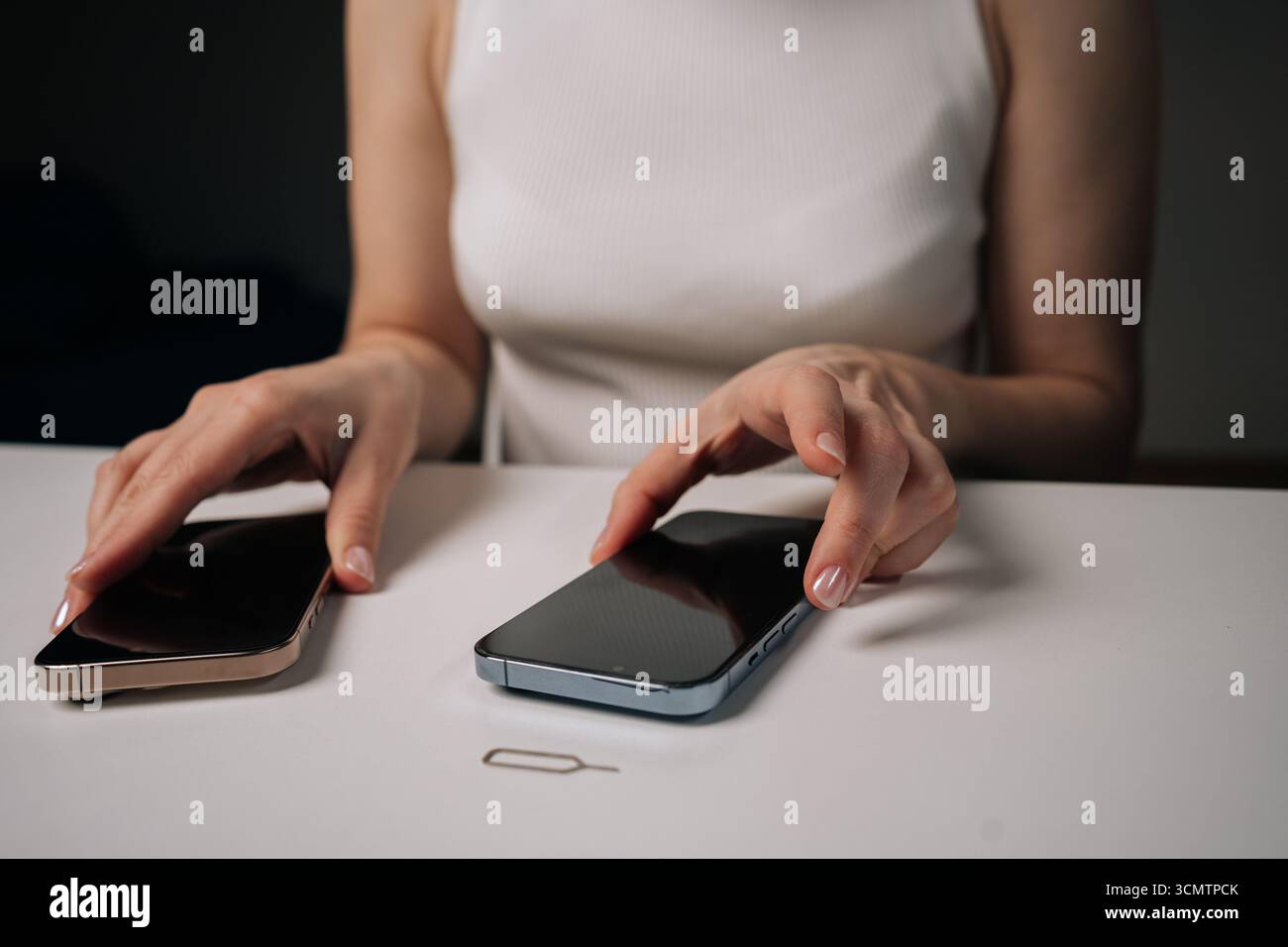 Frau, die altes Smartphone mit neuem Mobiltelefon vergleicht, Funktionen und Design genau untersucht und das sim-Auswurfwerkzeug in der Nähe auf einem weißen Tisch platziert. Stockfoto