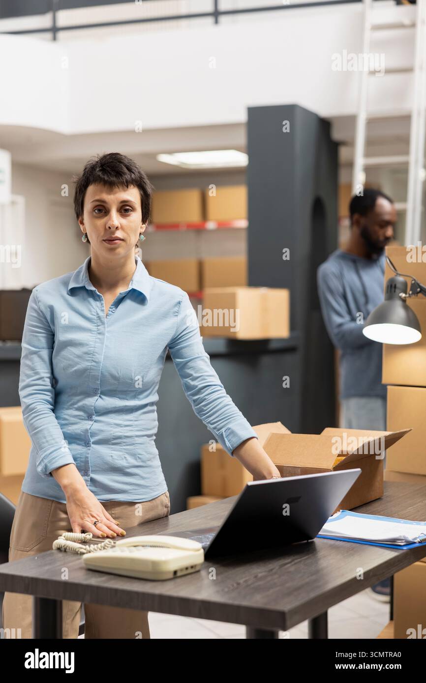 Eine junge Frau an ihrem Schreibtisch überprüft Sendungsdaten für Lieferdienste und überprüft Kassetten in einem Lagerraum, bevor sie zur Verteilung geschickt wird. Handverpackte Waren für den Einzelhandel. Stockfoto