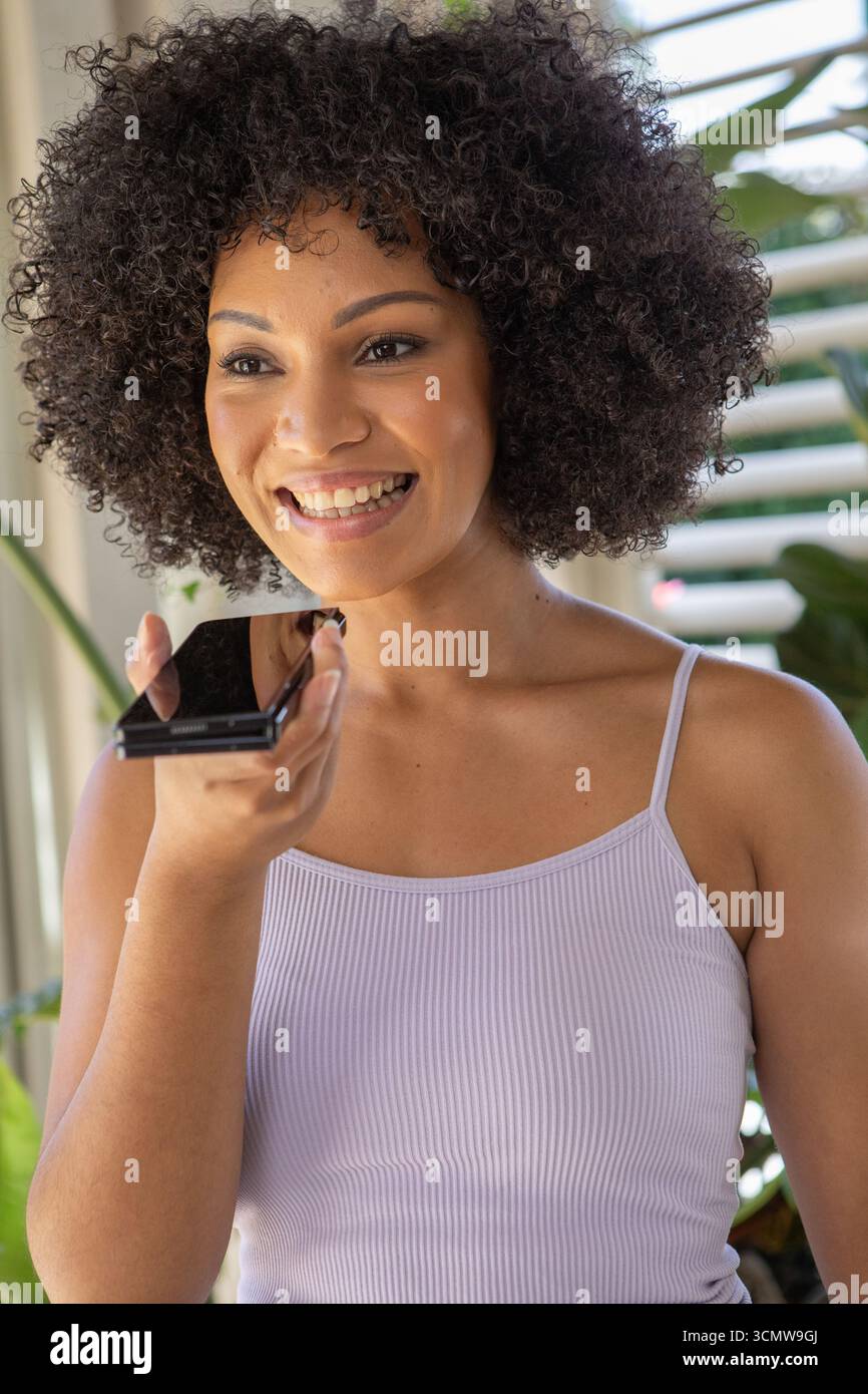 Erwachsene afroamerikanische Frau, die das Smartphone am Mund hält und zu Hause mit Pflanzen am Fenster spricht Stockfoto