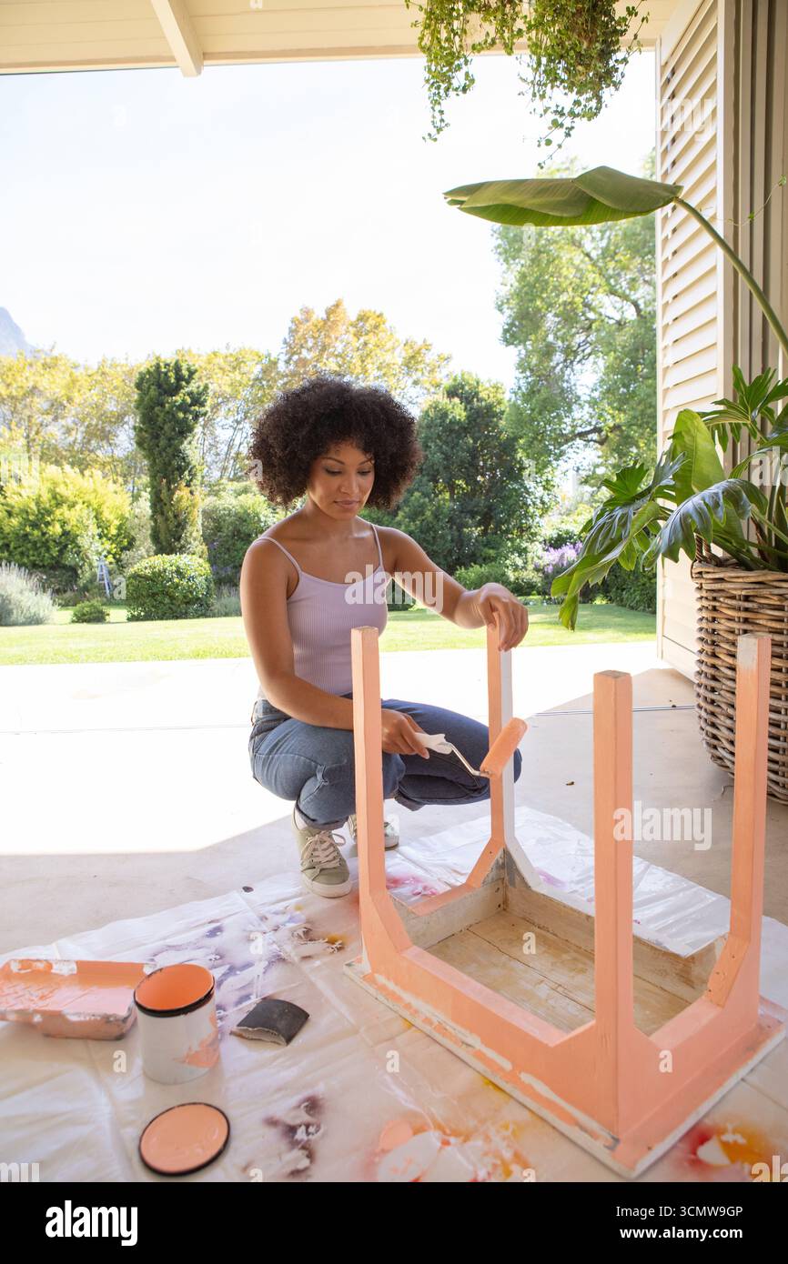afroamerikanerin, die Pfirsichfarbe auf der Unterseite des Seitentischs mit Roller auf der Terrasse aufträgt Stockfoto