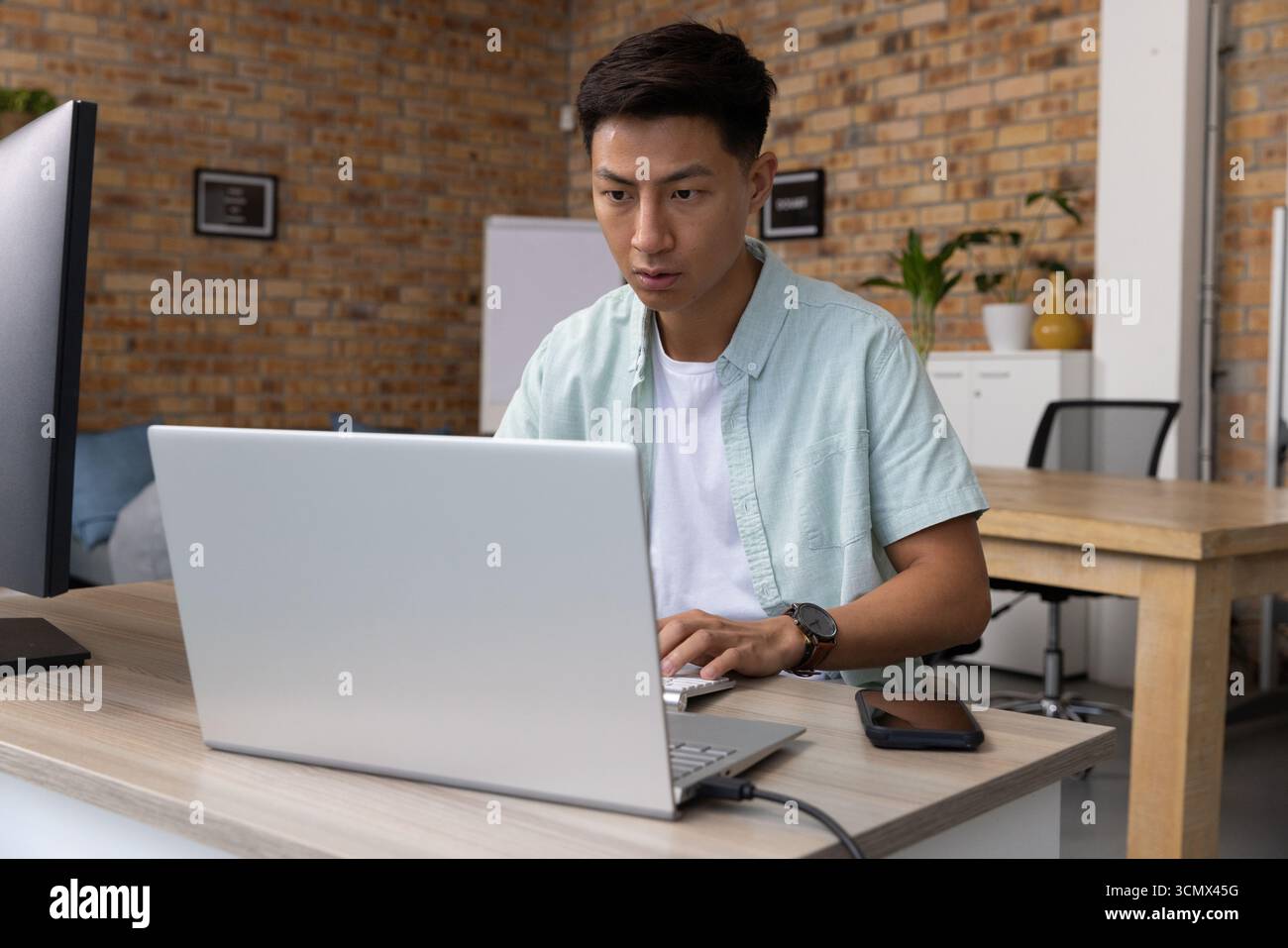 Asiatischer Mann, der auf silbernem Laptop am Schreibtisch im Backsteinstudio mit Monitor-Smartphone-Whiteboard tippt Stockfoto