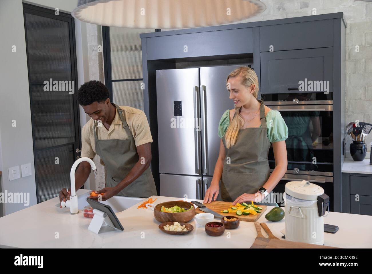 Diverse Paare waschen Karotten unter dem Wasserhahn, schneiden Zucchini auf Schneidebrett auf der Kücheninsel Stockfoto