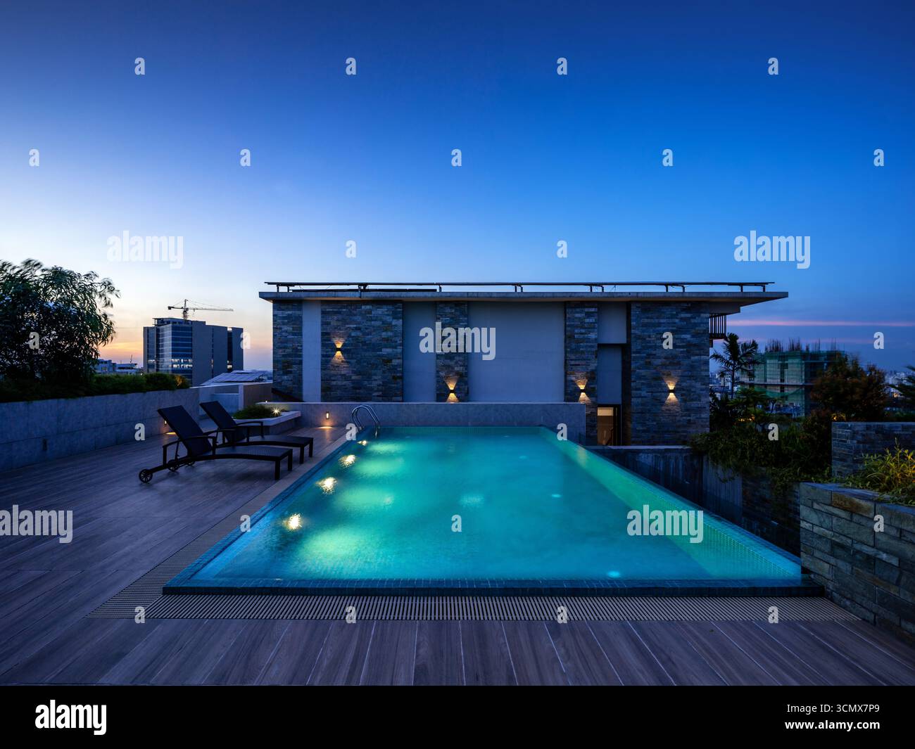 Moderner Swimmingpool auf dem Dach mit Liegestühlen und gedämpfter Beleuchtung in der Abenddämmerung. Zeitgenössische Architektur und ruhige Atmosphäre schaffen eine luxuriöse Stadt. Stockfoto