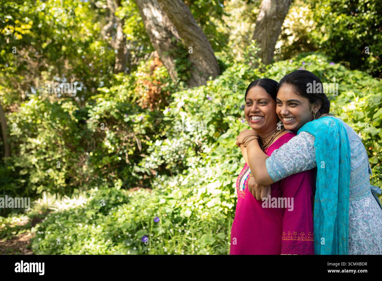 Indische Mutter und Tochter umarmen und lächeln im üppigen Garten mit farbenfrohen Kurtas, Kopierraum Stockfoto