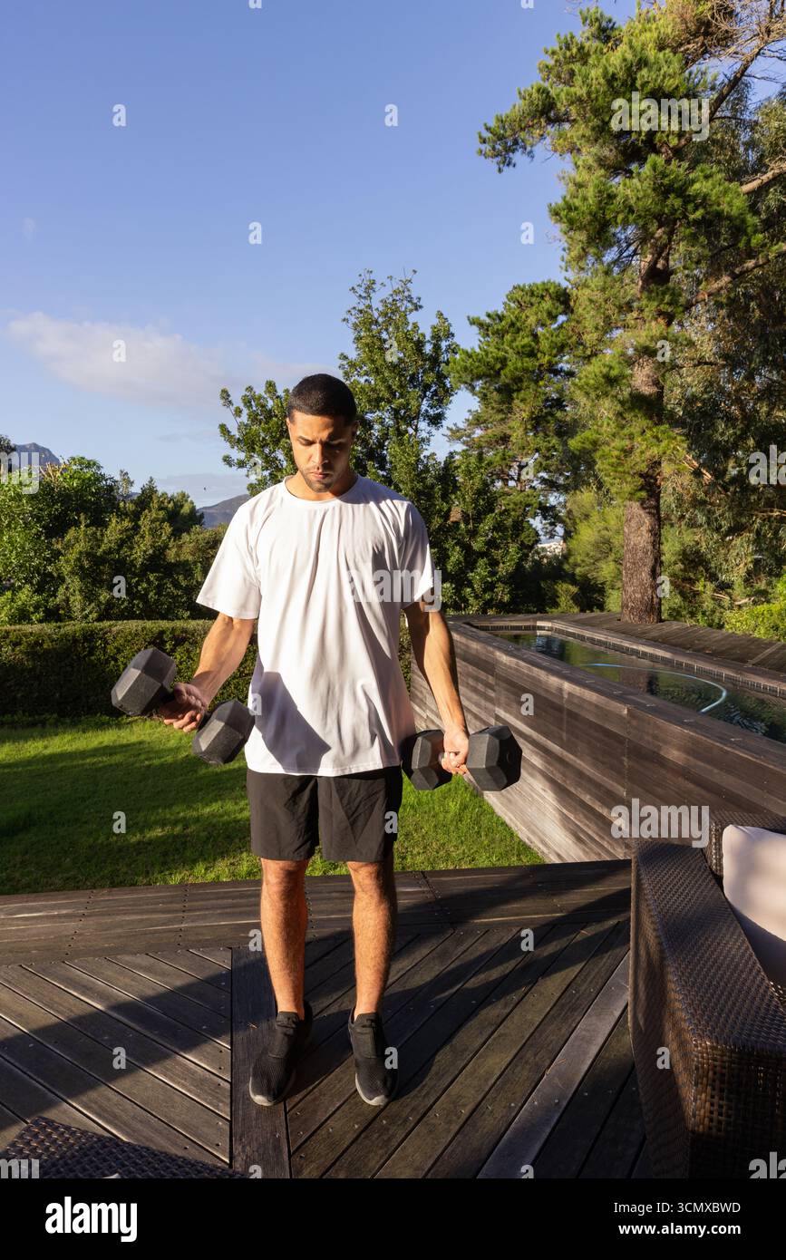 Hispanischer Erwachsener, männlich mit Trainingsausrüstung, Hanteln auf Deck neben dem Pool, Korbsessel Stockfoto