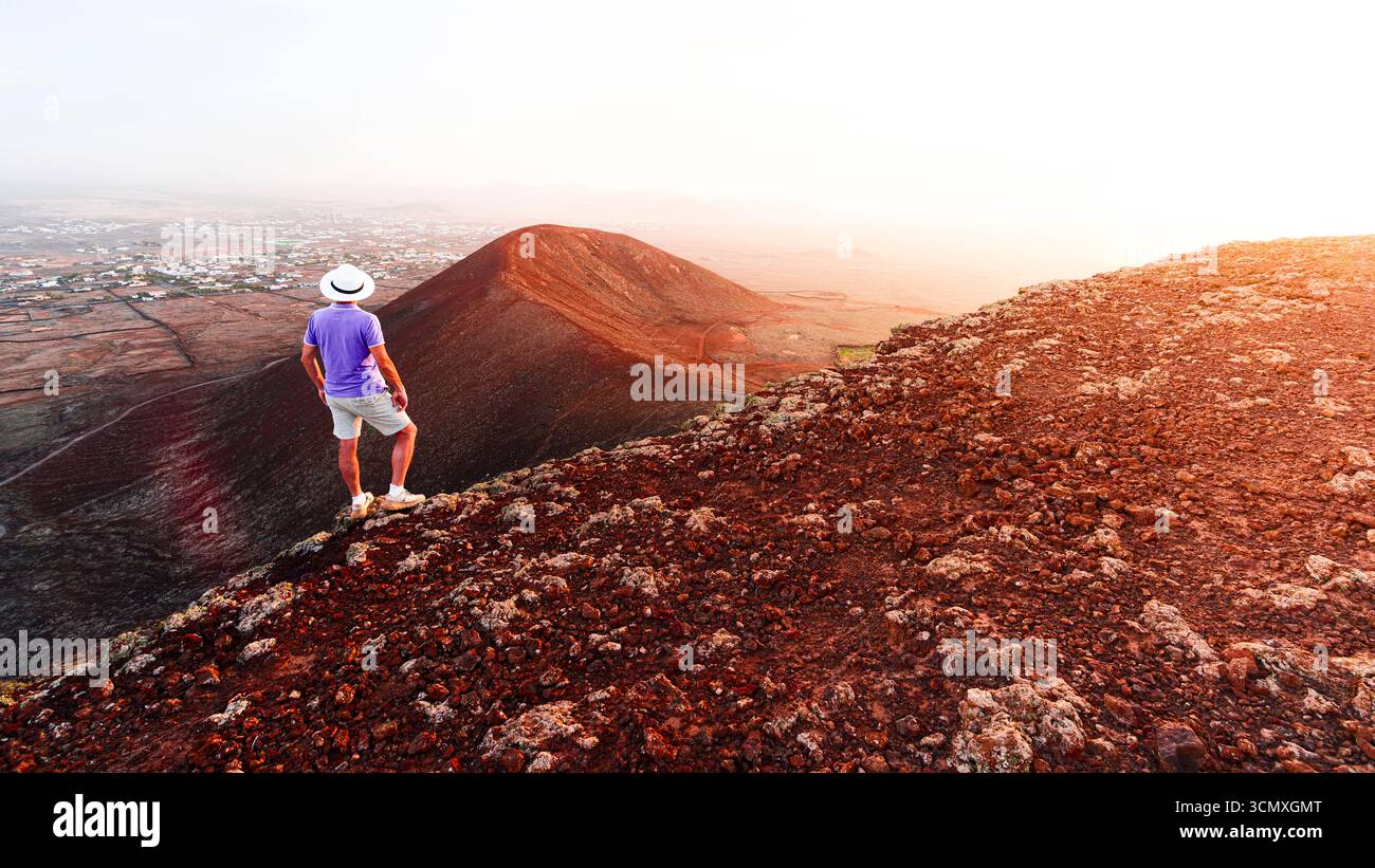 Rückansicht eines Mannes mit lässiger Kleidung und Hut, der den Sonnenuntergang auf dem Vulkan Calderon Hondo, Fuerteventura, den Kanarischen Inseln, Spanien beobachtet Stockfoto