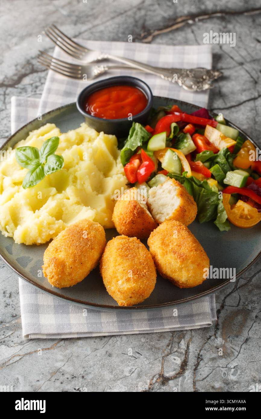 Nahaufnahme von spanischen Hähnchenkroketten mit Kartoffelpüree, Gemüsesalat und Ketchup auf einem Teller auf einem Tisch. Vertikal Stockfoto