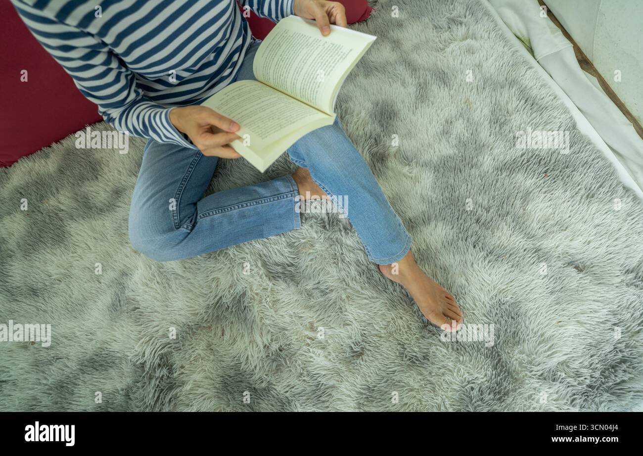 Eine Frau, die barfuß auf einem flauschigen Teppich sitzt, liest ein Buch in lässigen Jeans und gestreiftem Hemd. Entspannung, Achtsamkeit und digitaler Detox-Lebensstil für Stockfoto