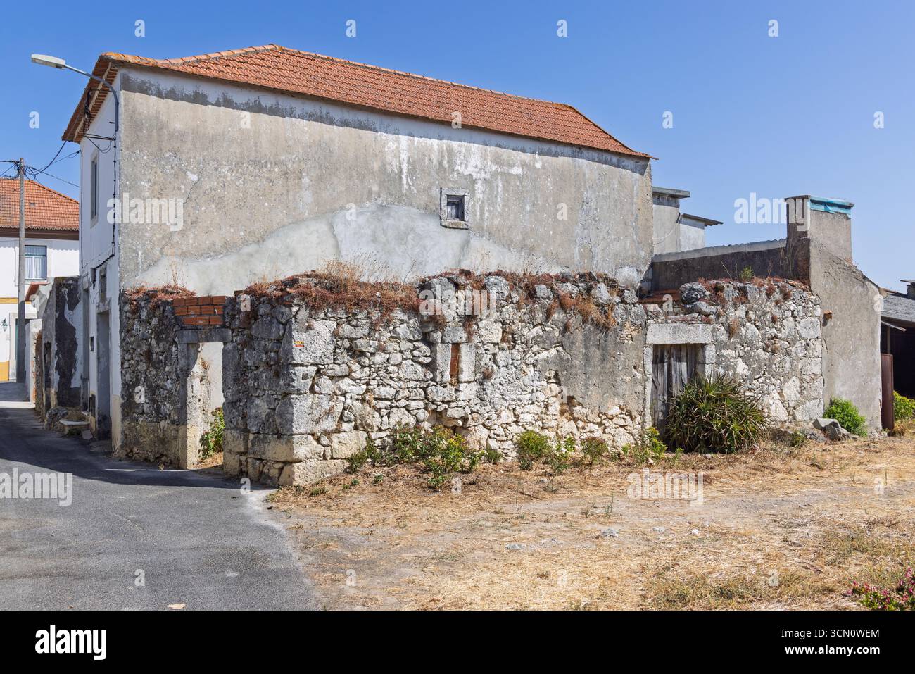 Ein Seitenblick auf ein verlassenes portugiesisches Gebäude mit zerbröckelnden Steinmauern, einem rot gekachelten Dach und trockenem Gras davor, das seinen Zustand der Vernachlässigung verdeutlicht Stockfoto