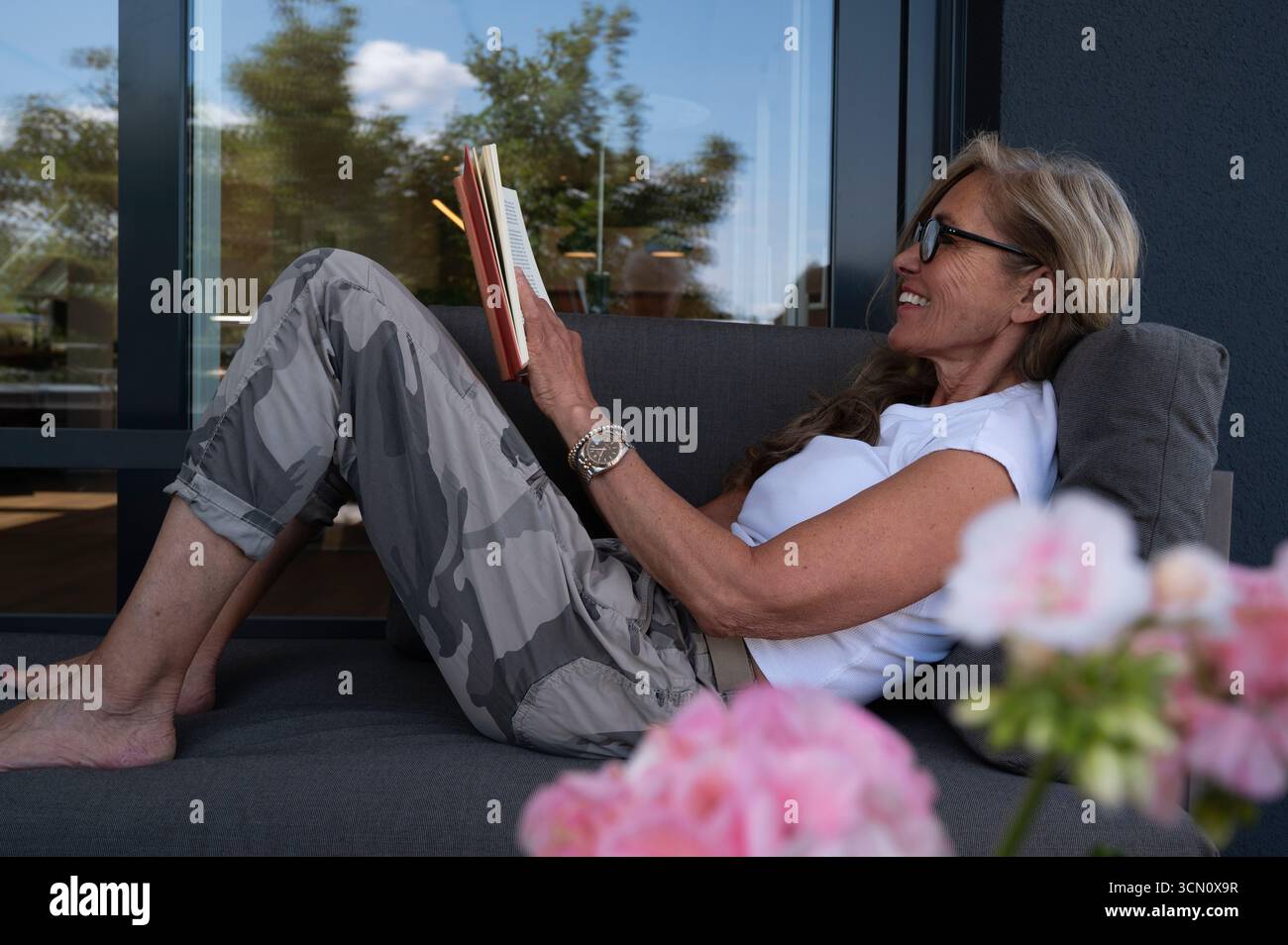 Eine ältere Frau mit Brille sitzt zu Hause auf dem Sofa auf der Terrasse und liest ein gutes Buch. Sie ist weitsichtig und genießt ihre Freizeit und ihren Ruhestand. Stockfoto