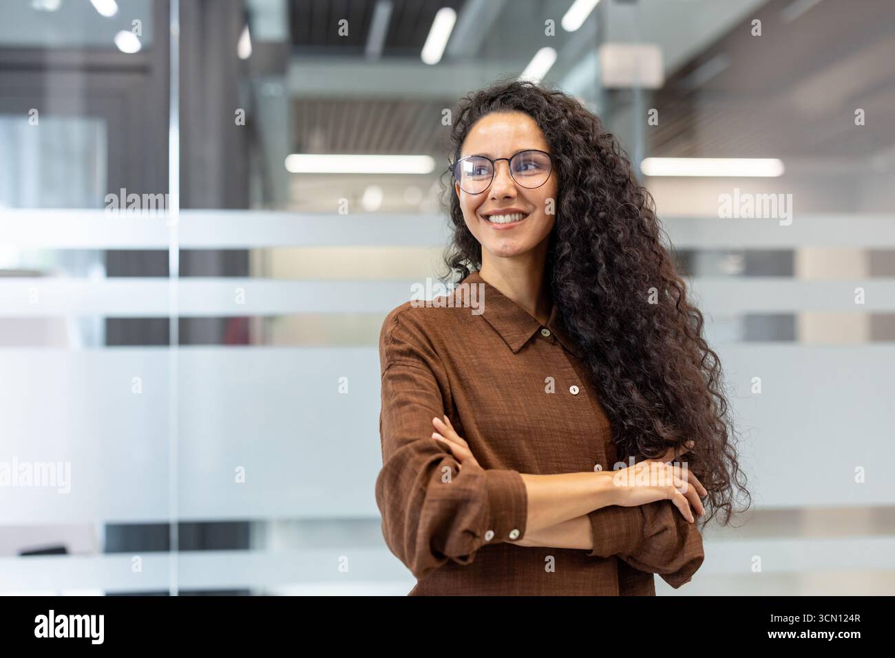 Selbstbewusste junge Frau mit lockigem Haar und Brille, die in einem modernen Büroumfeld steht, die Arme kreuzt und wegblickt, steht für Erfolg und Professionalität im Geschäft Stockfoto